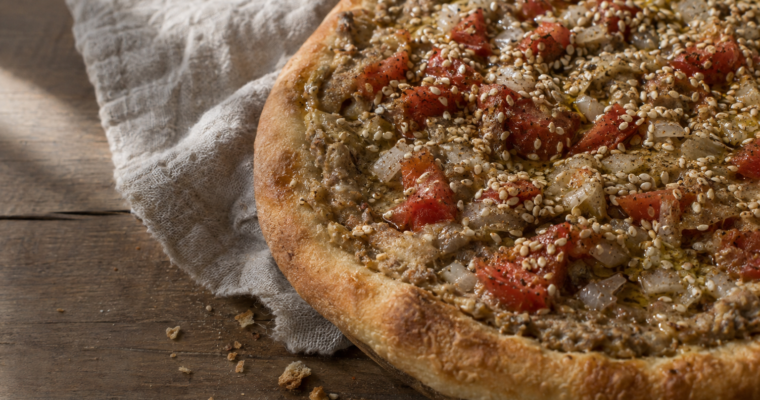 Una focaccia manaqish bi kishk con condimento di yogurt, pomodoro e cipolla, servita su un tavolo di legno.