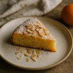 Una fetta di pan d'arancio decorata con zucchero a velo e lamelle di mandorle tostate, su un piatto di ceramica, accanto a un'arancia non trattata.